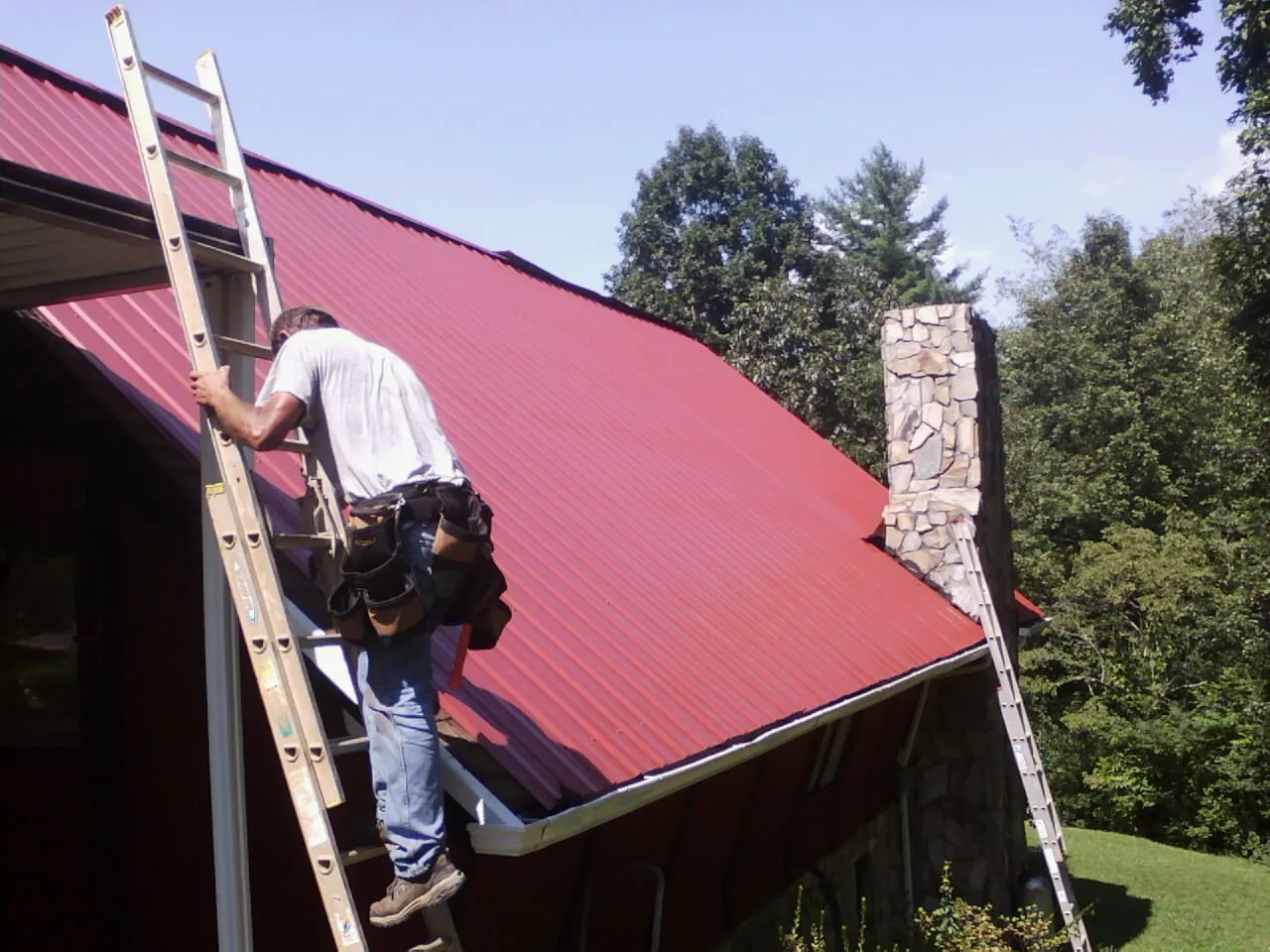 Roofer on a ladder beside a red metal roof and stone chimney — Soffit & Fascia Repair by Roofing Brothers in Hillister